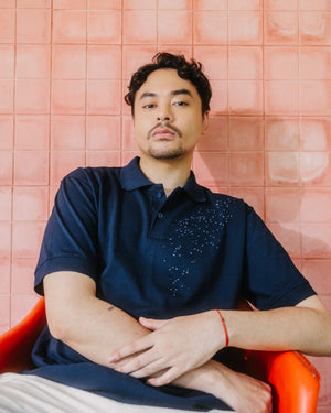 A man wearing a navy blue polo shirt with a decorative pattern on the chest, sitting in an orange chair against a pink tiled wall.