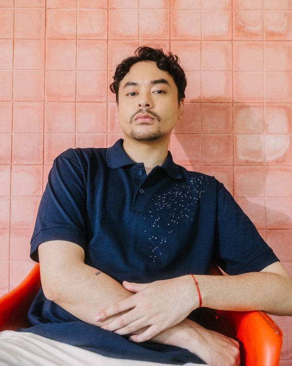 A man wearing a navy blue polo shirt with a decorative pattern on the chest, sitting in an orange chair against a pink tiled wall.
