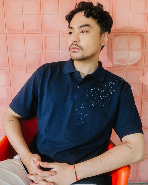 A man wearing a navy blue polo shirt with a small white embroidered pattern on the chest, sitting in an orange chair against a pink tiled wall.
