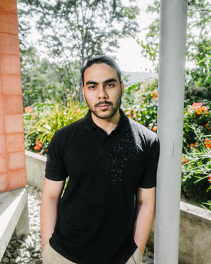 A man wearing a black short-sleeve polo shirt with a small white speckled graphic on the chest, standing outdoors next to a concrete pillar with green foliage and flowers in the background.