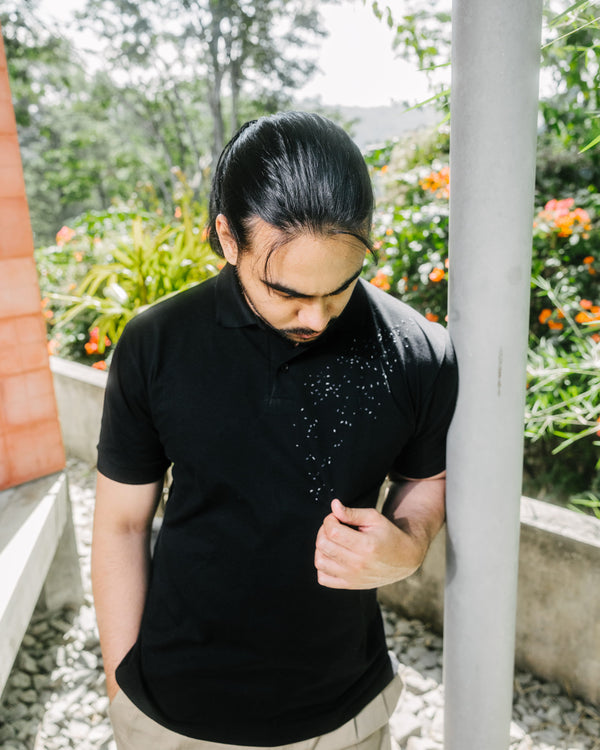 A man with long dark hair tied back wears a black short-sleeve polo shirt with a small white speckled graphic on the chest, standing outdoors next to a white pillar with green foliage and flowers in the background.